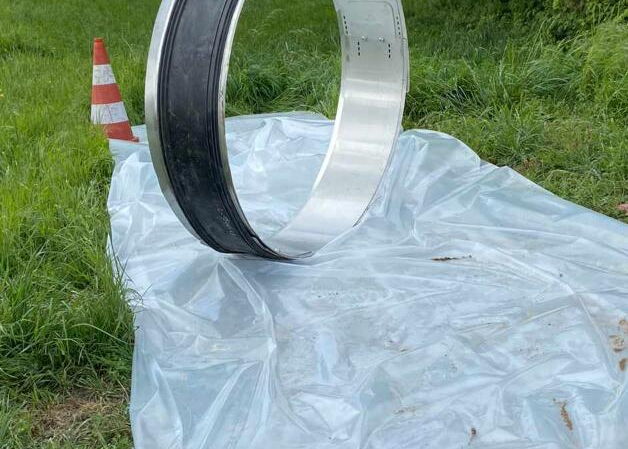 Großer Metallring steht auf einer Plastikplane neben einem Grasweg, im Hintergrund ein Verkehrskegel.