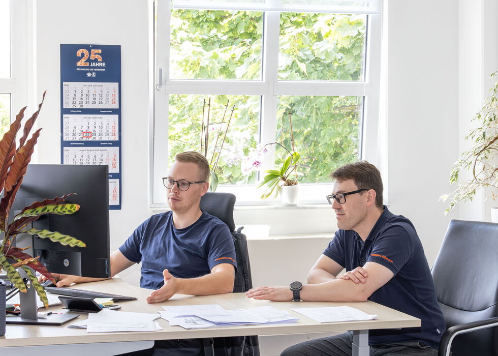 Zwei Männer sitzen an einem Schreibtisch in einem hellen Büro und besprechen Unterlagen.