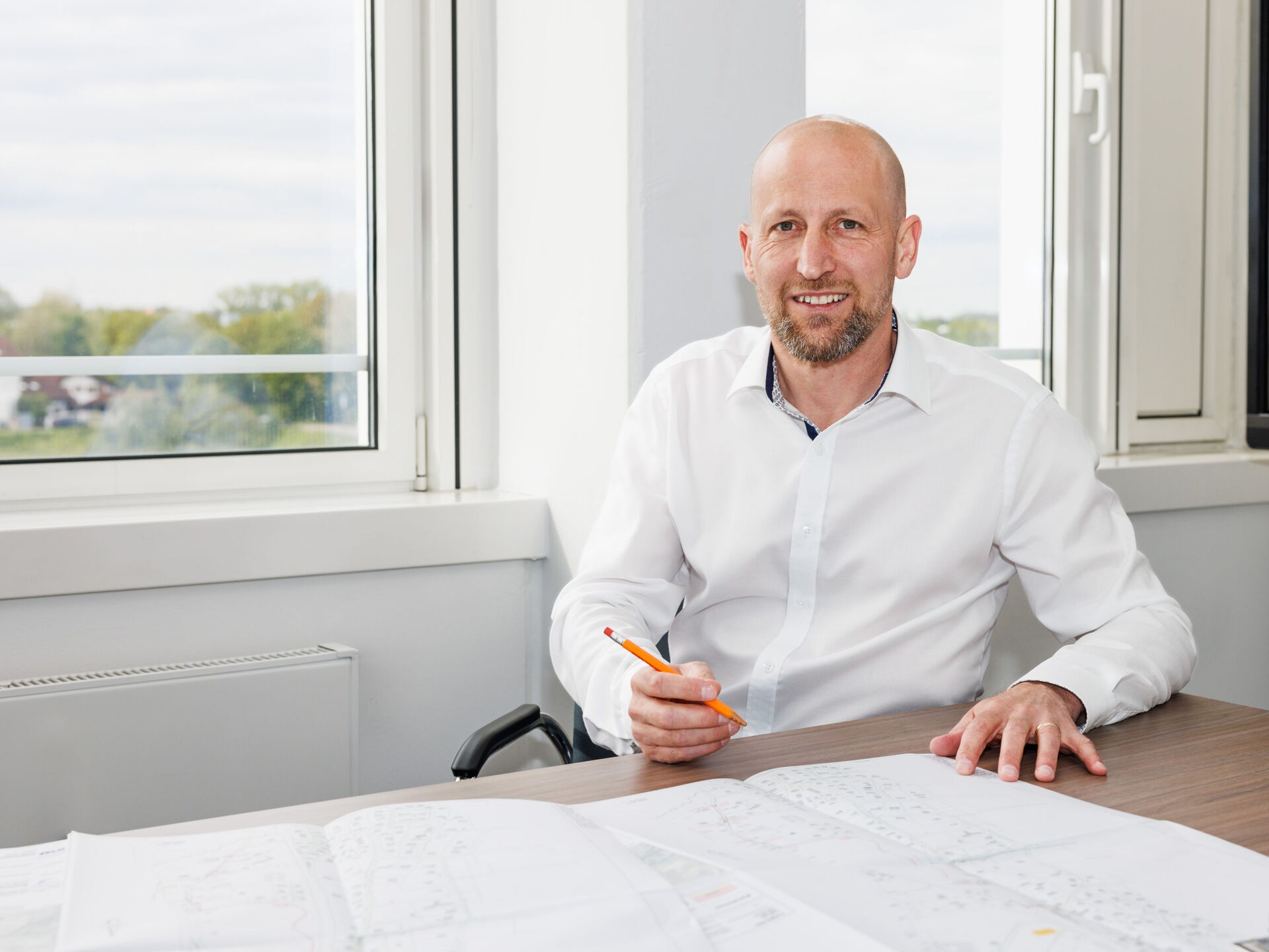 Der lächelnde Dipl.-Ing. Peter Prossliner im weißen Hemd sitzt mit Bleistift an einem Schreibtisch neben großen Bauplänen am Fenster.