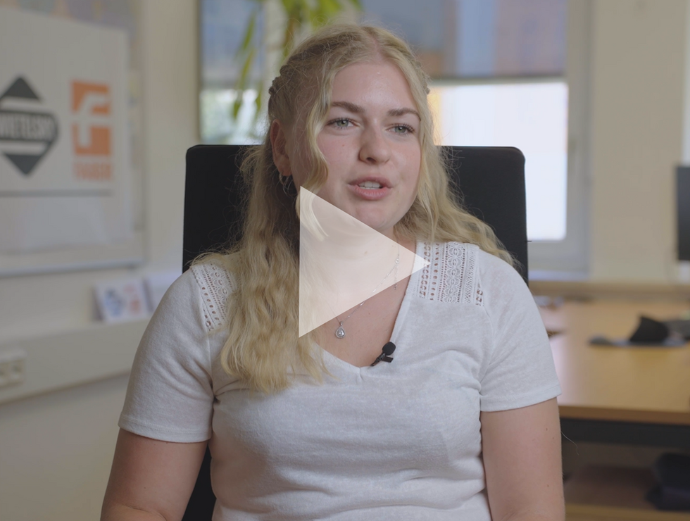 Junge Frau mit blonden Haaren spricht in einem Büro, im Hintergrund Schreibtisch und Computer