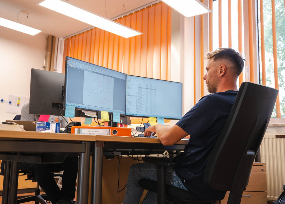 Ein Mann sitzt auf einem Bürostuhl an einem Schreibtisch mit zwei Monitoren in einem modernen Arbeitsraum.
