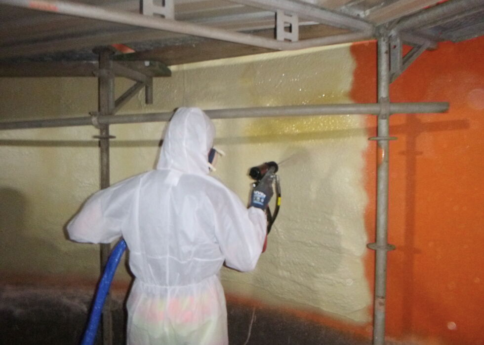 Arbeiter in Schutzanzug sprüht Beschichtung auf eine Wand im Stahlbehälter.