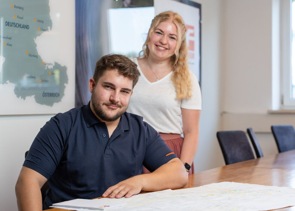 Zwei Kollegen posieren in einem hellen Büro mit Karte an der Wand und Konferenztisch im Vordergrund.