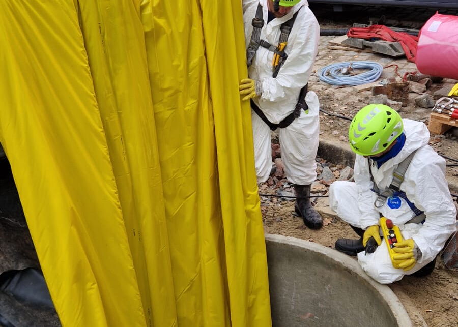 Techniker in Schutzanzügen arbeiten neben einem Betonbrunnen mit Sicherheitsausrüstung und Schläuchen.
