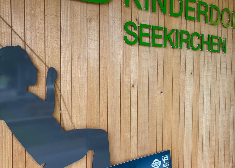 Grünes SOS-Kinderdorf-Seekirchen-Schild an einer Holzwand mit blauer Silhouette eines sitzenden Kindes und einem Infoposter darunter.