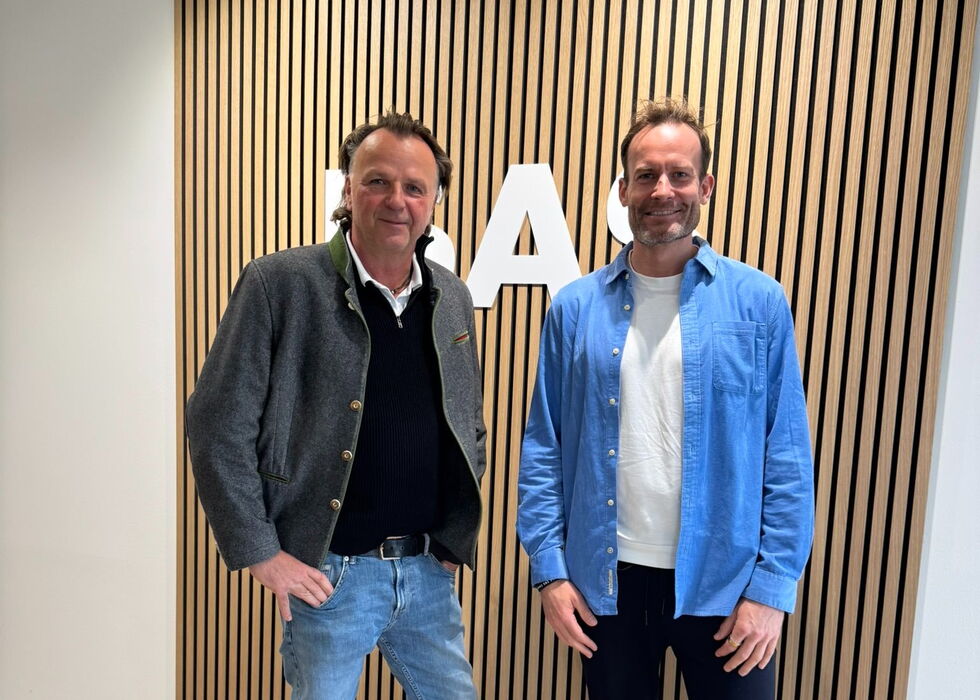 Jörg Brunecker und Alexander Jung posieren in einem Innenraum vor einer gestreiften Holzwand mit der Aufschrift ISAS.