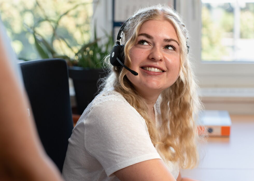 Eine lächelnde Frau mit Headset im Büro, die nach oben schaut und mit jemandem außerhalb des Bildes spricht.