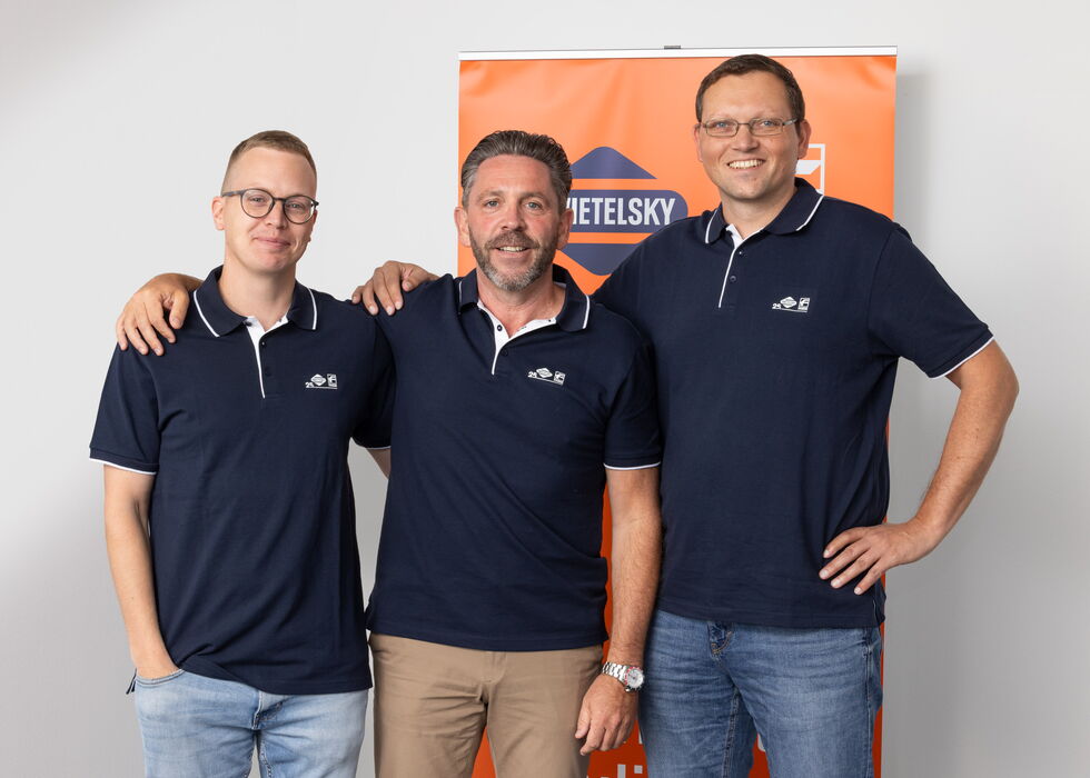Drei Männer in dunkelblauen Poloshirts stehen zusammen und lächeln vor einem orangefarbenen Banner.