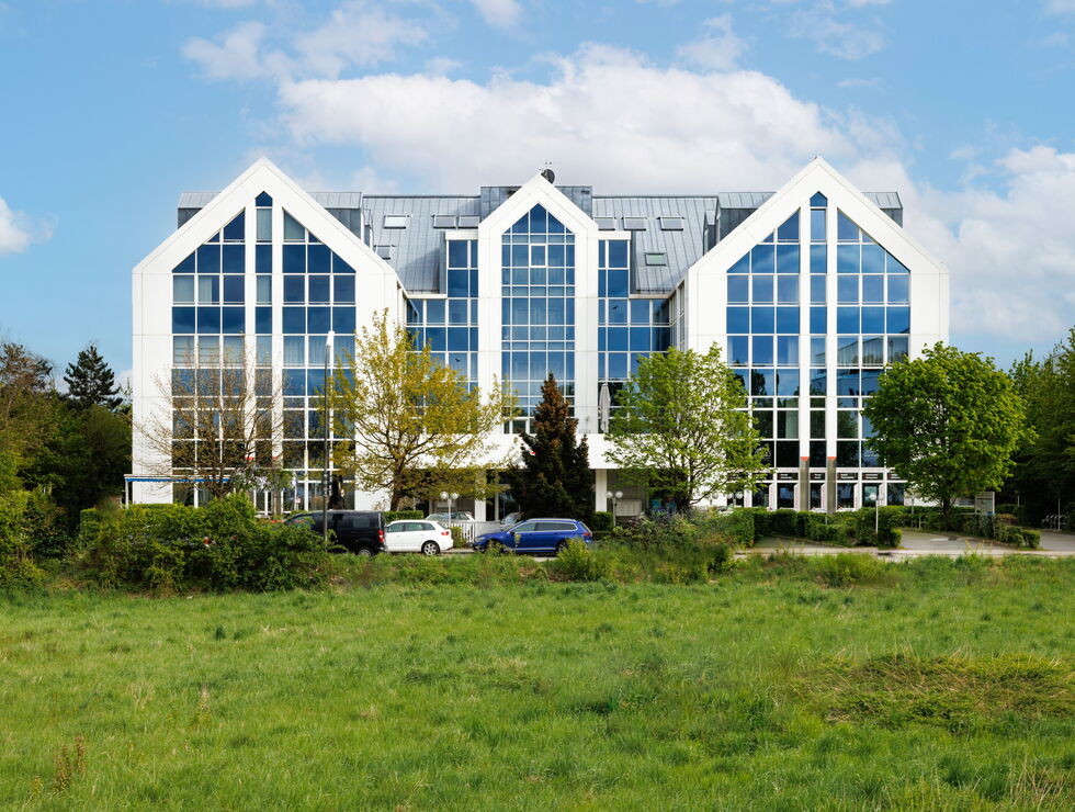 Ein modernes Bürogebäude mit großen Glasfenstern und weißen Giebelrahmen neben einer Wiese und Bäumen.