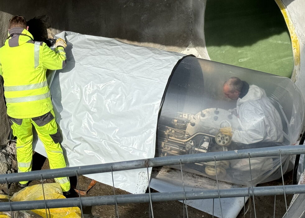 Ein Arbeiter in Warnkleidung steht neben einer mit Folie abgedeckten Maschine, in der eine weitere Person arbeitet.