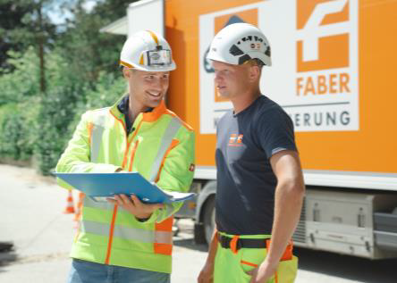 Zwei Arbeiter mit Schutzhelmen besprechen Pläne neben einem Swietelsky-Faber Lastwagen, einer hält ein Klemmbrett.