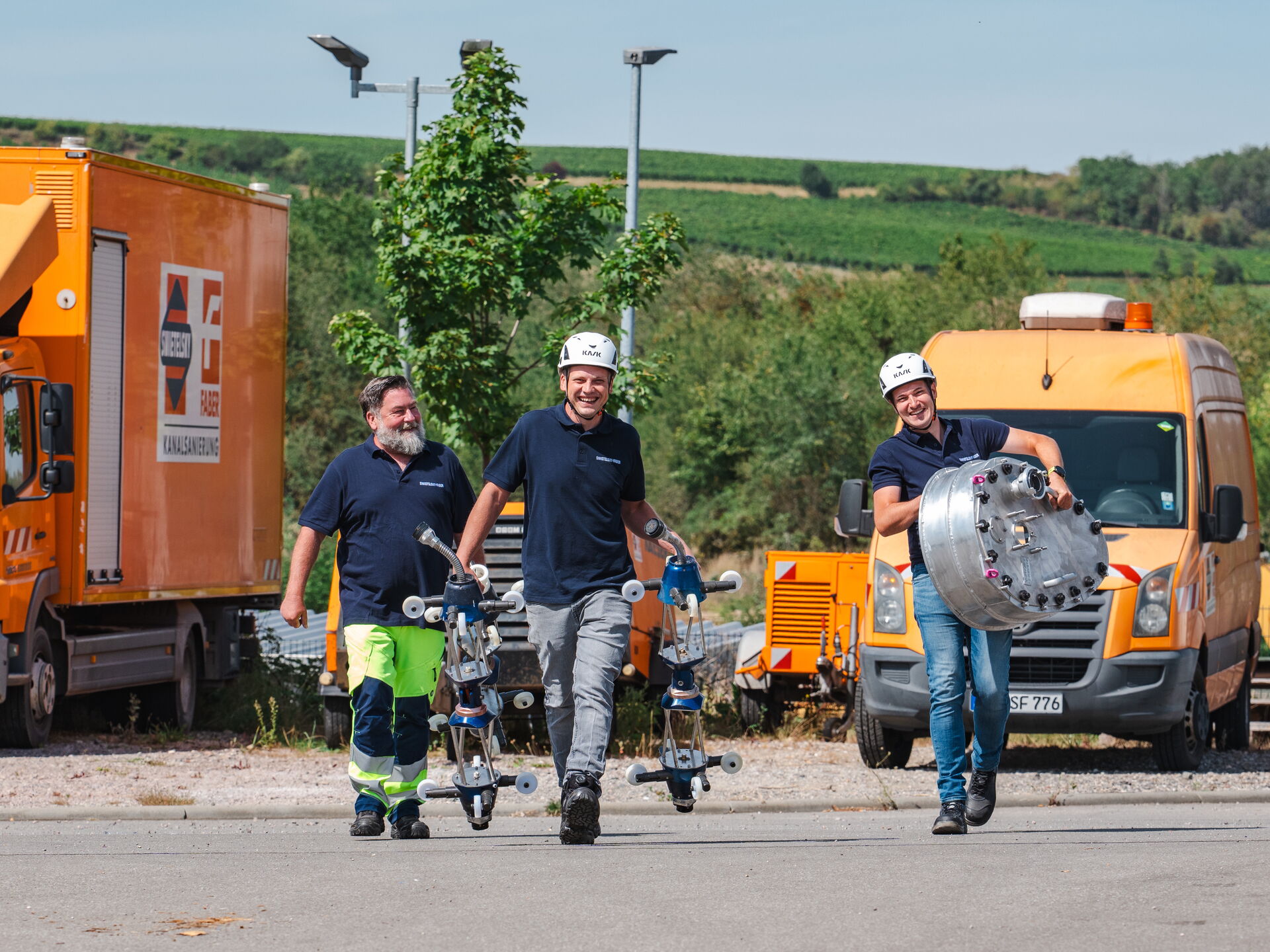 Drei Arbeiter mit Helmen tragen technische Geräte an einem sonnigen Tag mit grünen Hügeln im Hintergrund