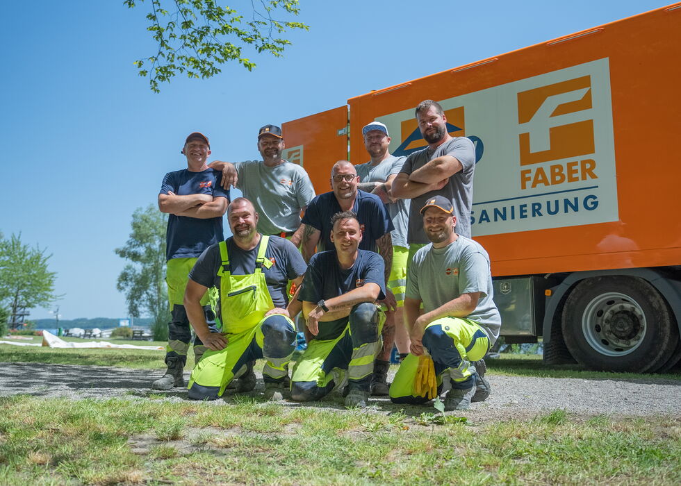 Sieben Bauarbeiter posieren und lächeln vor einem orangen FABER-LKW an einem sonnigen Tag