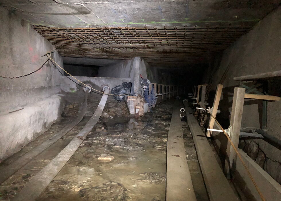 Schwach beleuchteter unterirdischer Tunnel mit Wasser auf dem Boden, Rohren rechts und verstreuten Baumaterialien