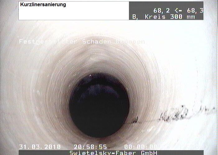 Kreisförmige Innenansicht eines Rohrs mit sichtbarer Beschädigung und Ablagerungen bei der Kanalinspektion.