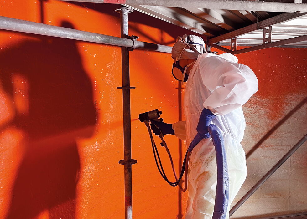 Ein Arbeiter im Schutzanzug sprüht unter einem Gerüst eine orange Wand.
