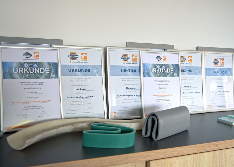 Framed certificates standing on a shelf beside rolled belts and sample materials.