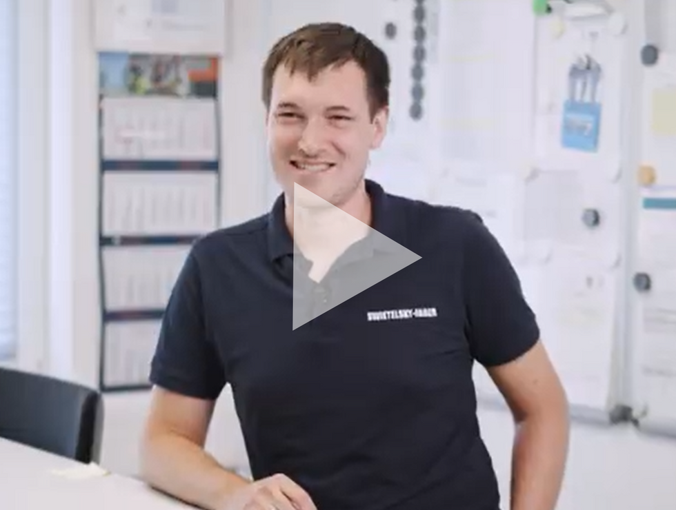 Ein lächelnder Mann in einem dunklen Poloshirt sitzt an einem Schreibtisch in einem hellen Büro.
