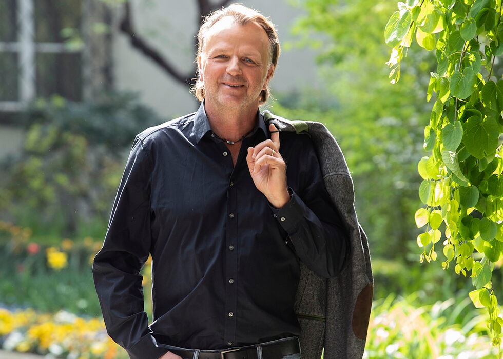 Jörg Brunecker in Jeans und dunklem Hemd geht einen Gartenweg entlang und trägt eine Jacke über der Schulter.