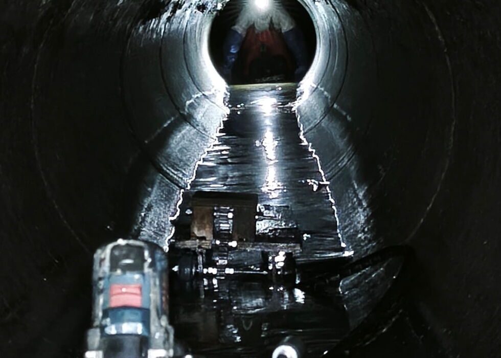 A pipe-laying system in a wet sewage tunnel, aligned with a well-lit opening.