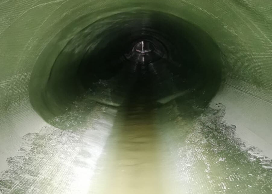 Blick in einen grünen zylindrischen Tunnel mit Wasser, das am Boden in eine dunkle Öffnung fließt.