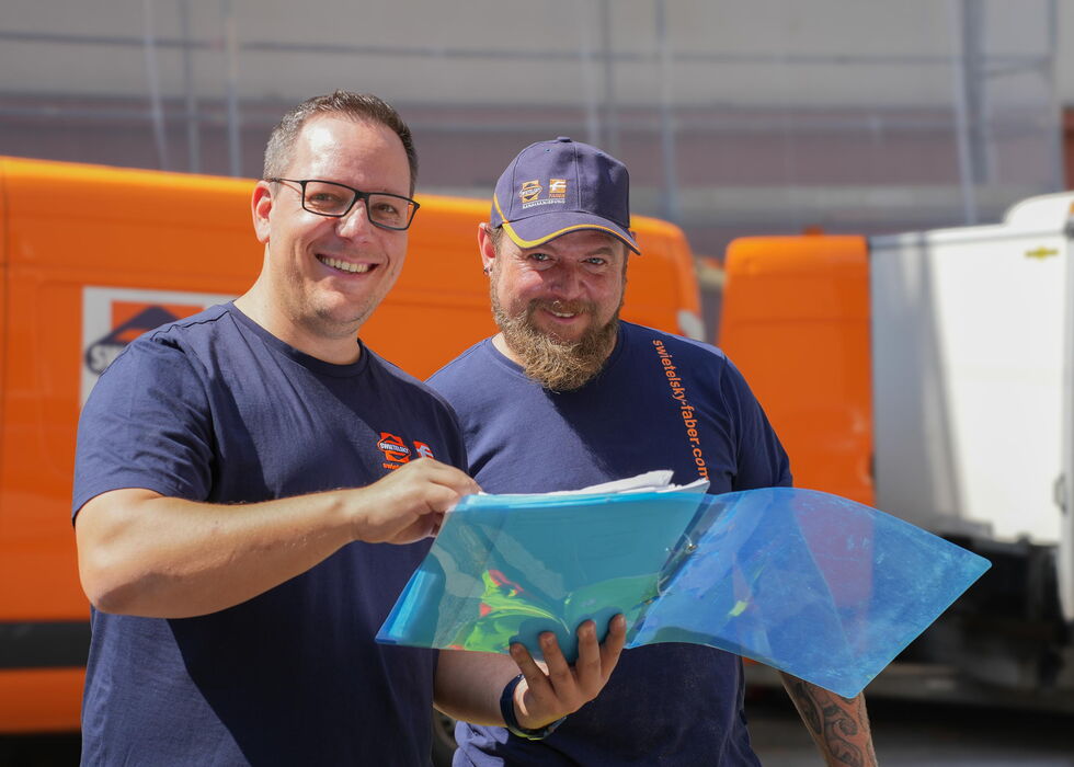 Zwei lächelnde Arbeiter vor einem orangefarbenen Fahrzeug prüfen gemeinsam einen Bauplan.
