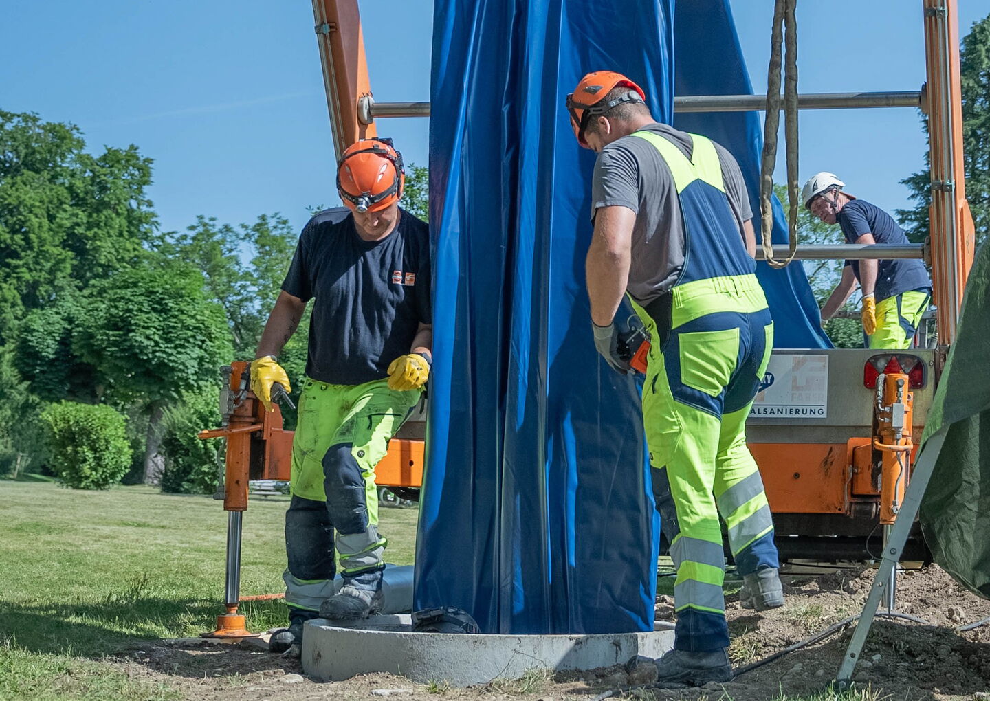 Arbeiter in Schutzkleidung prüfen im Freien eine große blaue Plane, die über Industrieanlagen hängt.