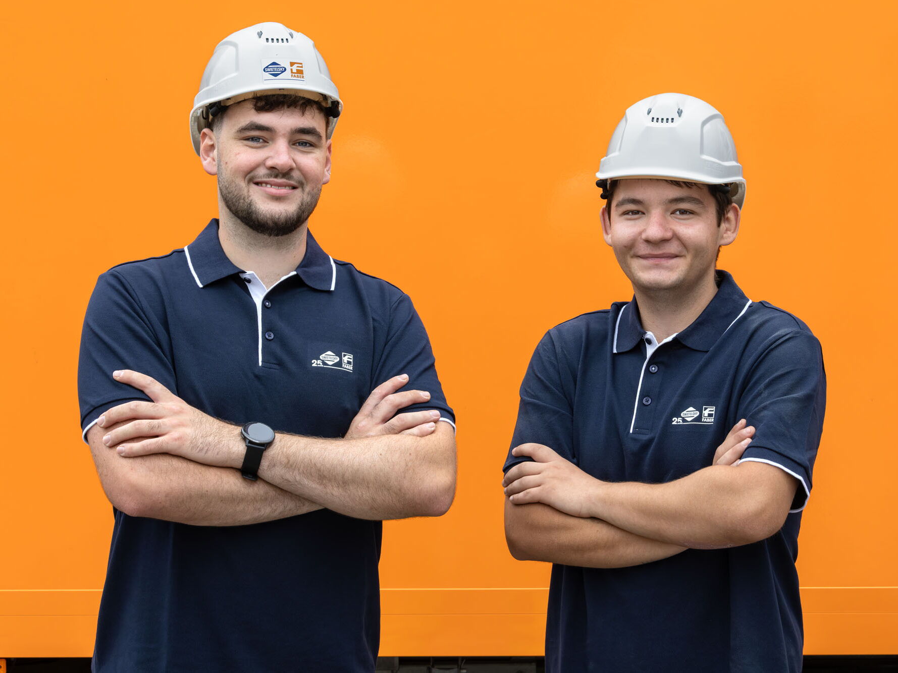 Zwei lächelnde Bauarbeiter mit Schutzhelmen und dunkelblauen Poloshirts stehen mit verschränkten Armen vor orangefarbenem Hintergrund.