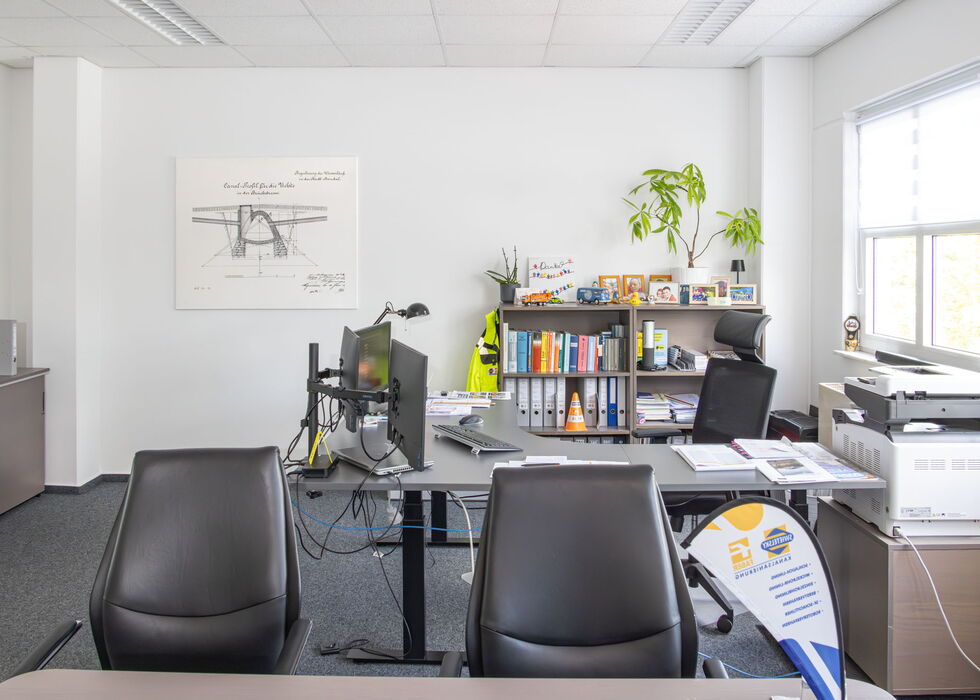 Helles modernes Büro mit Glastisch, zwei schwarzen Stühlen, Bücherregal und Fenster.