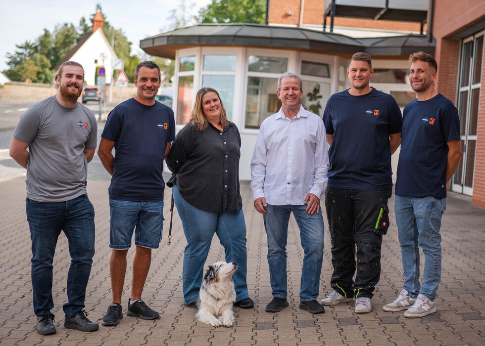 Sechs Personen stehen draußen vor einem Gebäude, mit einem kleinen Hund vorne.