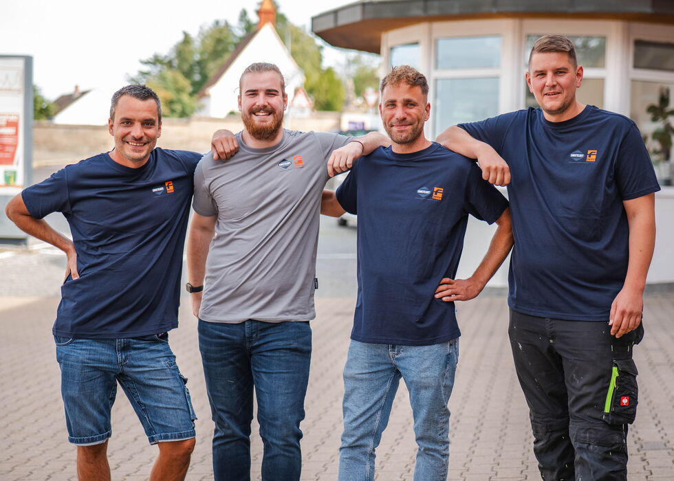 Vier Männer in Markenshirts stehen lächelnd zusammen vor einem Gebäude.