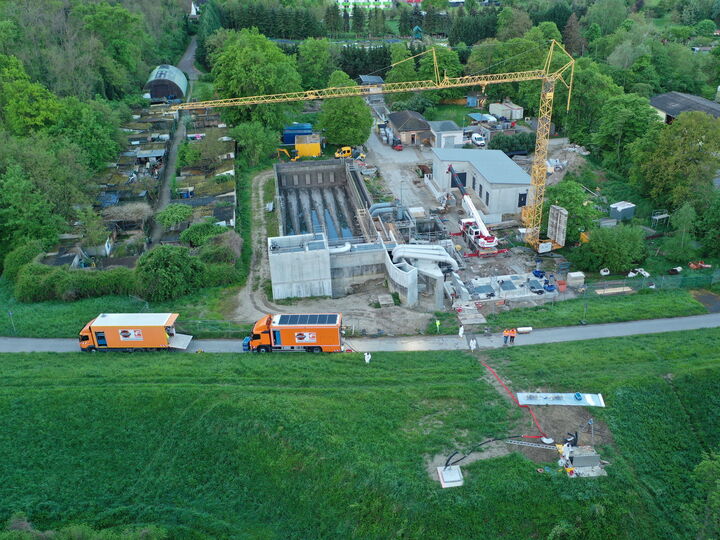 Luftaufnahme einer Baustelle mit Kran, im Bau befindlichen Gebäuden und Lastwagen neben einer Straße, umgeben von Feldern und Bäumen.