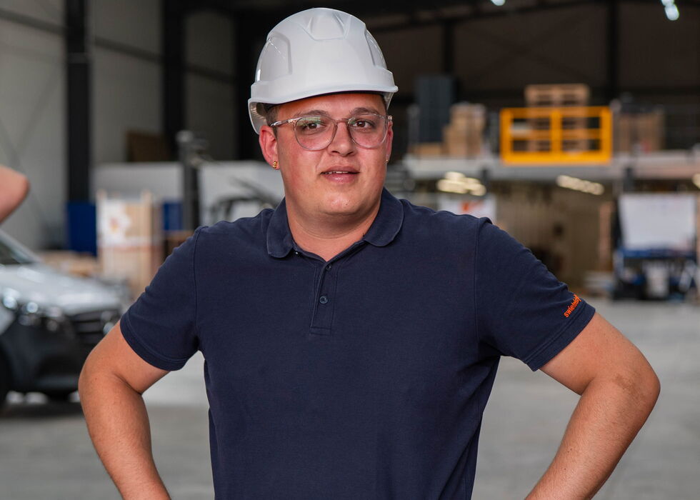 Ein Mann mit Schutzhelm steht mit den Händen in die Hüften gestützt in einer Lagerhalle.