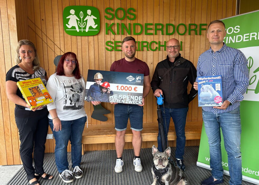 Fünf Personen stehen in einem Innenraum vor einem SOS-Kinderdorf-Schild und halten einen Spendencheck und Bücher; ein Hund sitzt vorn.