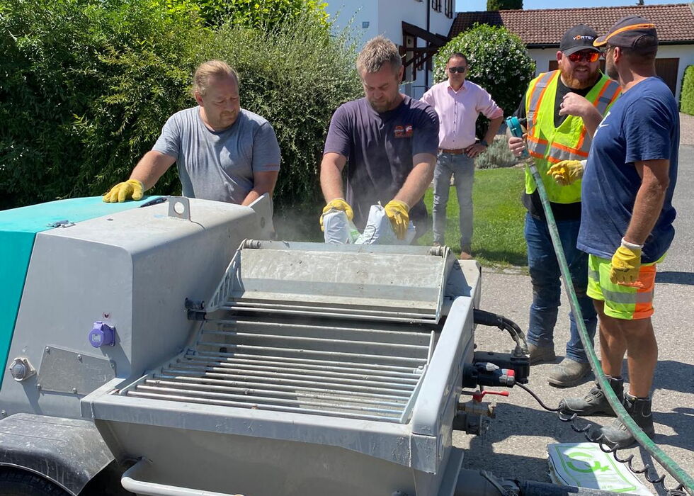 Mehrere Arbeiter mit Handschuhen und Warnwesten reinigen an einer sonnigen Wohnstraße eine mobile Kehrmaschine.