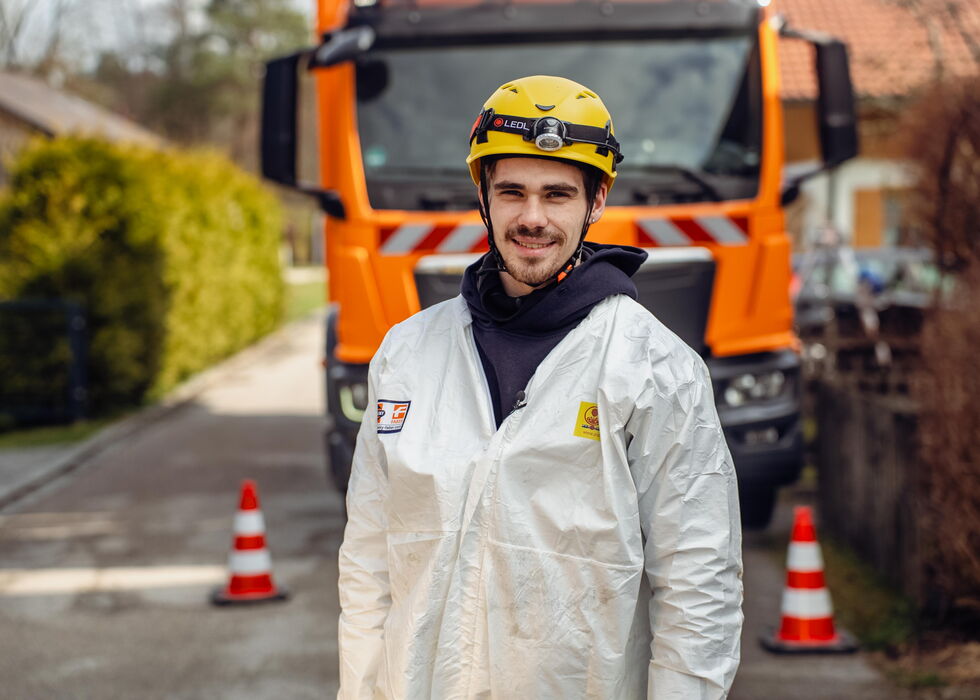 Lächelnder Straßenarbeiter mit Helm und weißem Schutzanzug vor einem orangefarbenen Nutzfahrzeug, mit Verkehrskegeln in der Nähe.