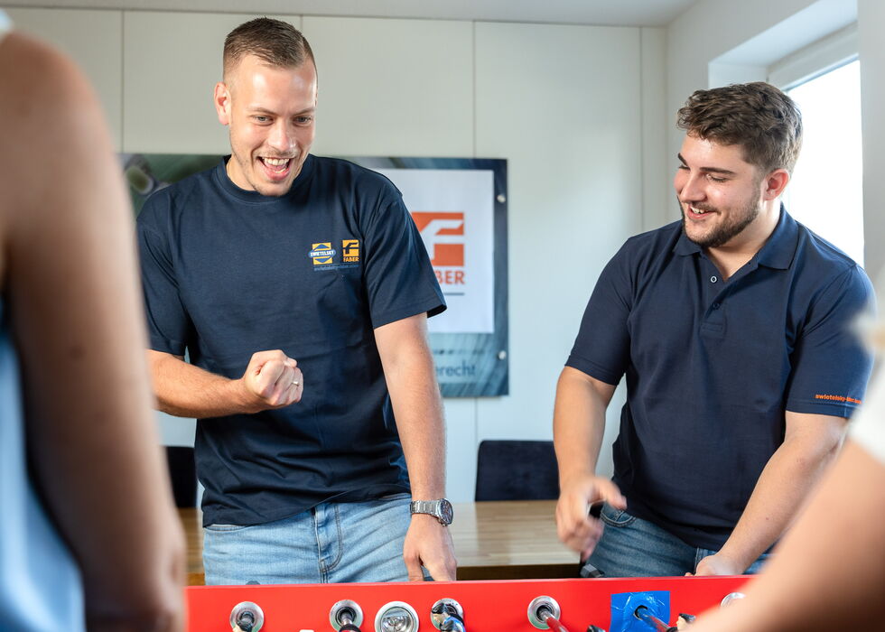 Zwei Männer lächeln und spielen Tischfußball in einem hellen Besprechungsraum.
