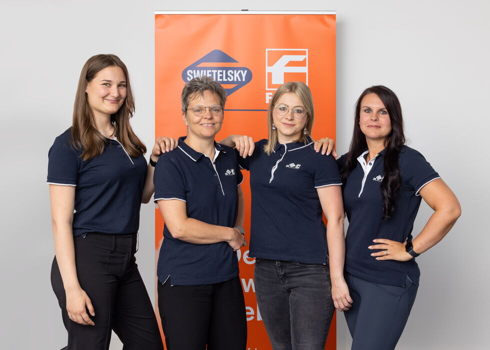 Vier Frauen stehen gemeinsam vor einem orangefarbenen Firmenbanner und lächeln in die Kamera.