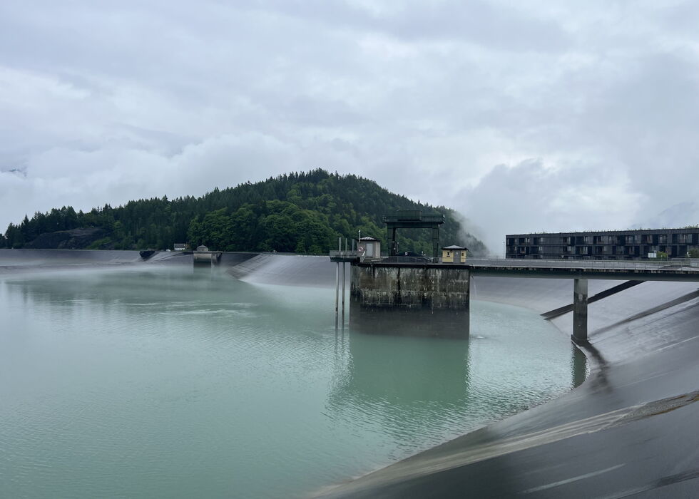 Ein Damm an einem ruhigen Fluss gibt Wasser ab; Gebäude und Bäume säumen das Ufer unter einem wolkigen Himmel.