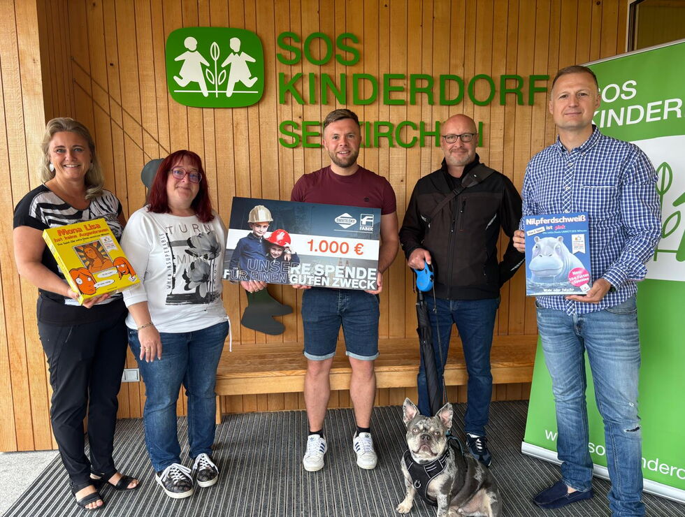 Fünf Personen stehen in einem Innenraum vor einem SOS-Kinderdorf-Schild und halten einen Spendencheck und Bücher; ein Hund sitzt vorn.