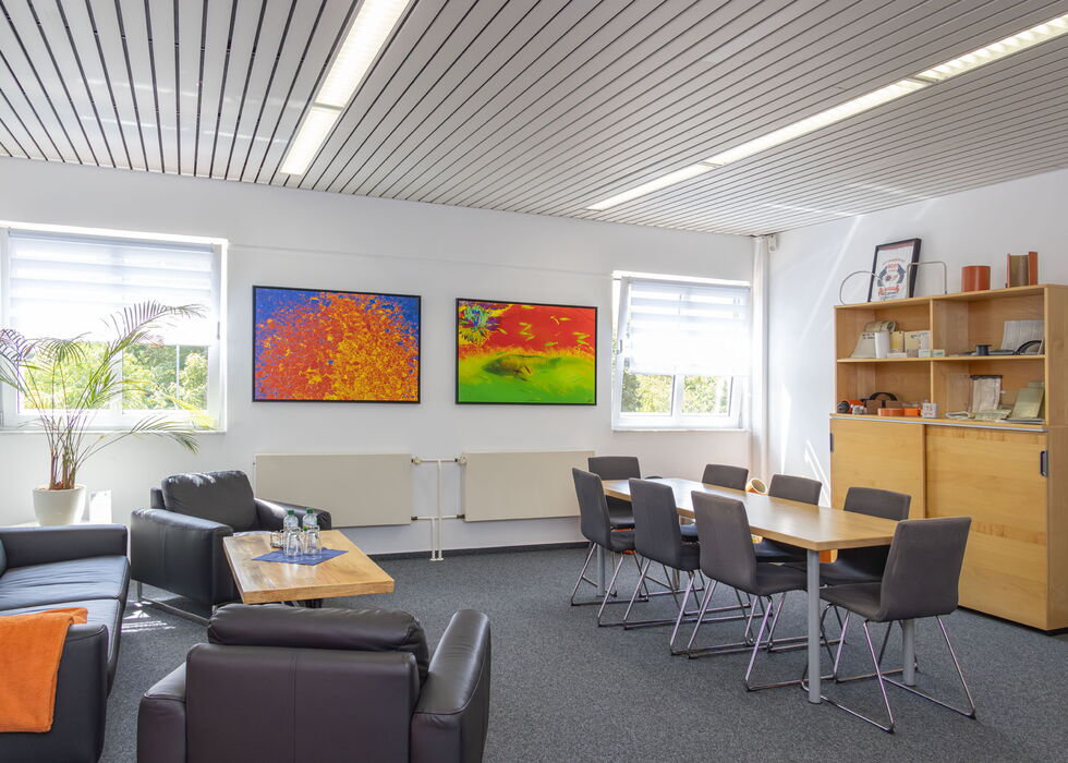 Heller Besprechungsraum mit Sofas, Tisch, Stühlen und bunten Bildern an der Wand.