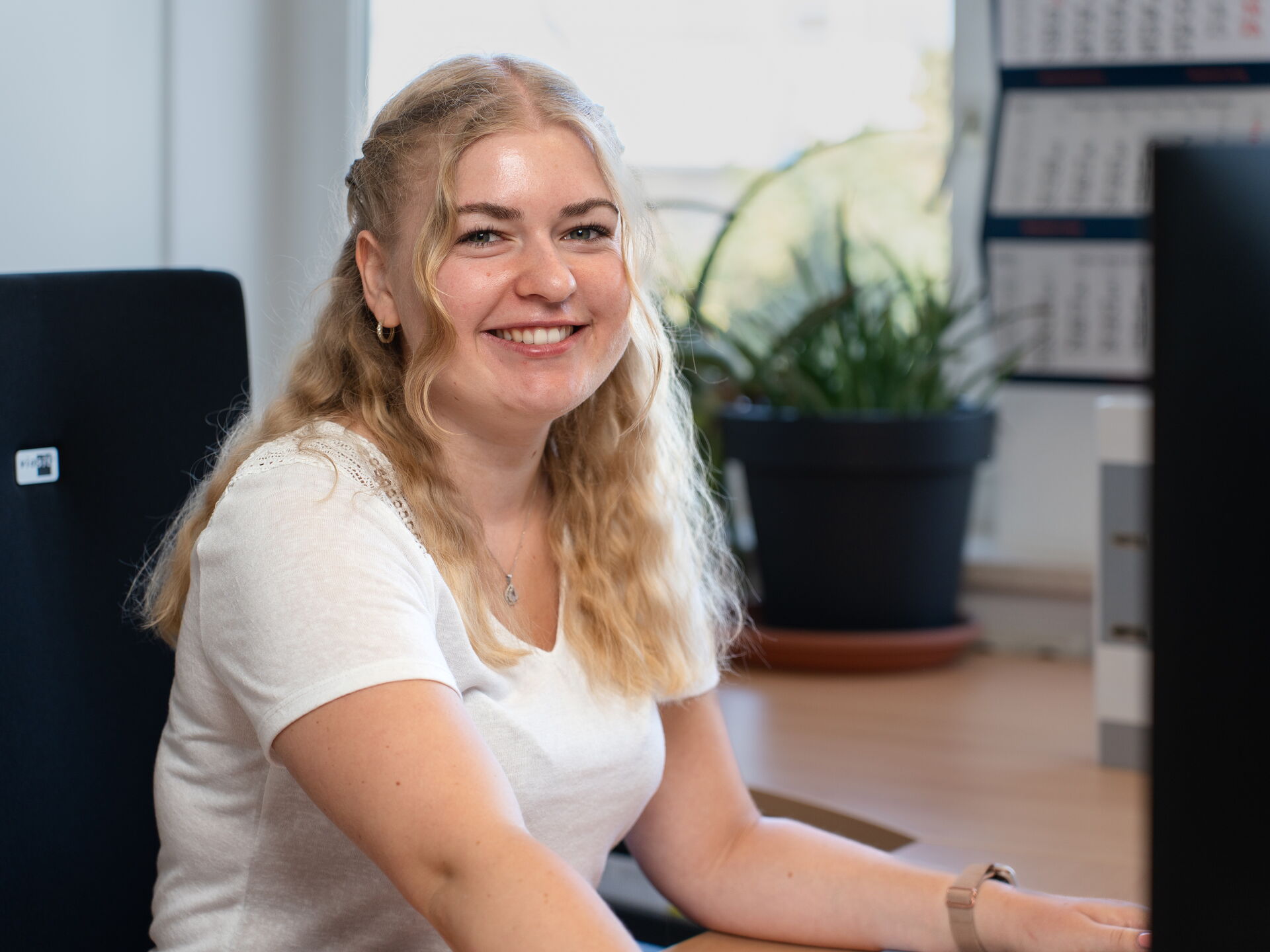 Junge Frau mit blonden Haaren sitzt auf einem Bürostuhl und lächelt, während sie am Computer arbeitet