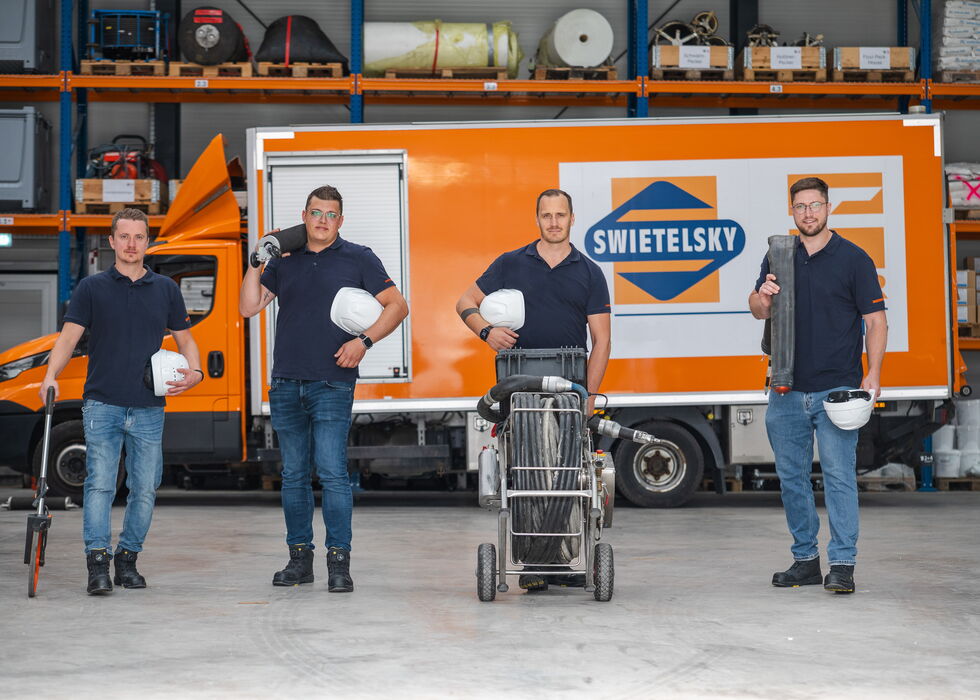 Vier Arbeiter mit Helmen stehen in einer Halle vor einem orangefarbenen SWIETELSKY-Lkw.