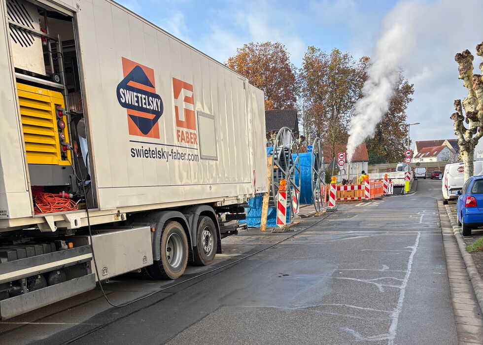 Ein Lkw steht an einer Straße neben einer Baustelle mit Absperrungen, Geräten und aufsteigendem Dampf in der Ferne.