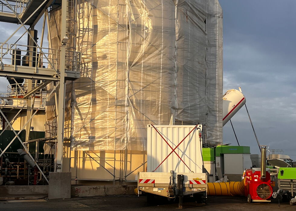 Ein großer Industriesilo ist mit Schutzfolie umhüllt, daneben stehen Ladegeräte in einer Anlage.