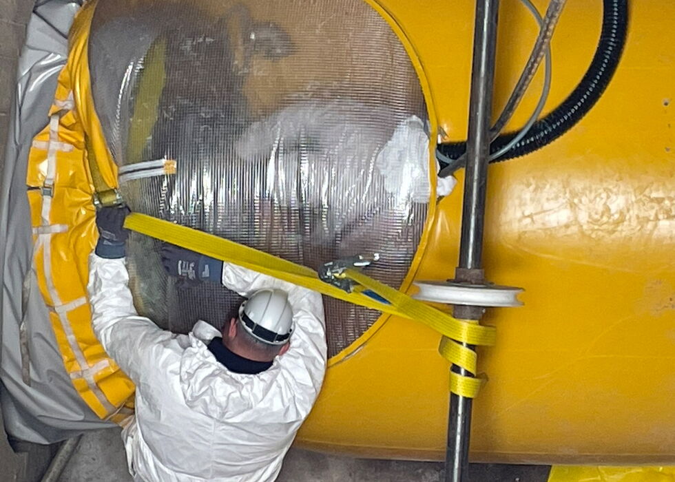 Mitarbeiter im Schutzanzug arbeitet an einem gelben Industriebottich mit Isolierung und Schläuchen in einer Werkstatt.