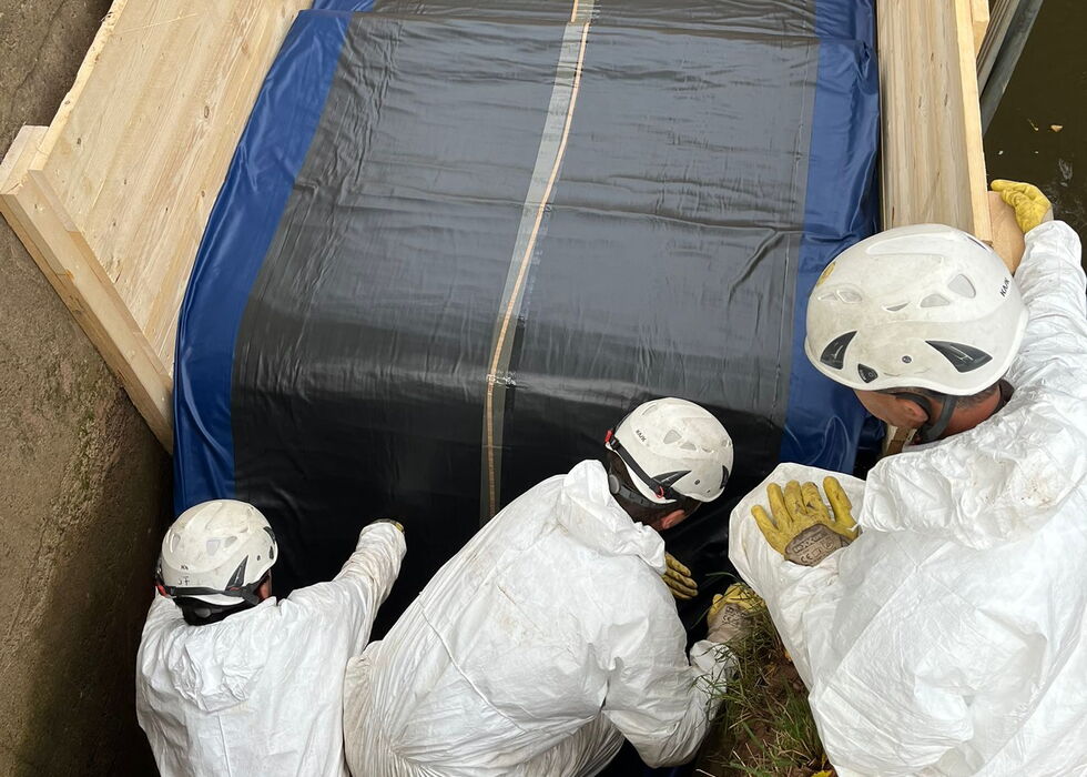 Forensiker in weißen Schutzanzügen untersuchen einen gesicherten Außenbereich mit blau abgedecktem Container und Abdeckfolie.