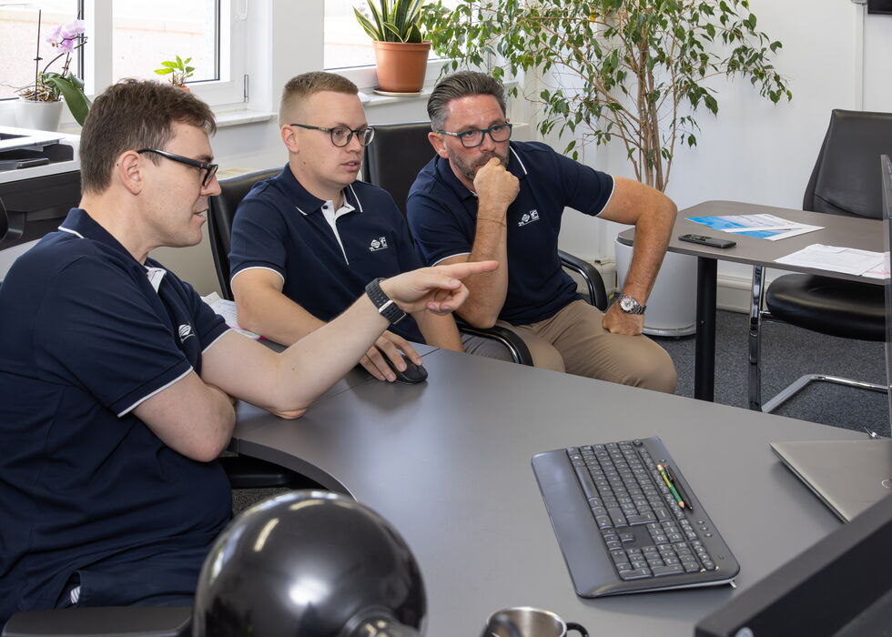 Drei Kollegen sitzen im Büro an einem Schreibtisch und besprechen etwas auf einem Computerbildschirm.