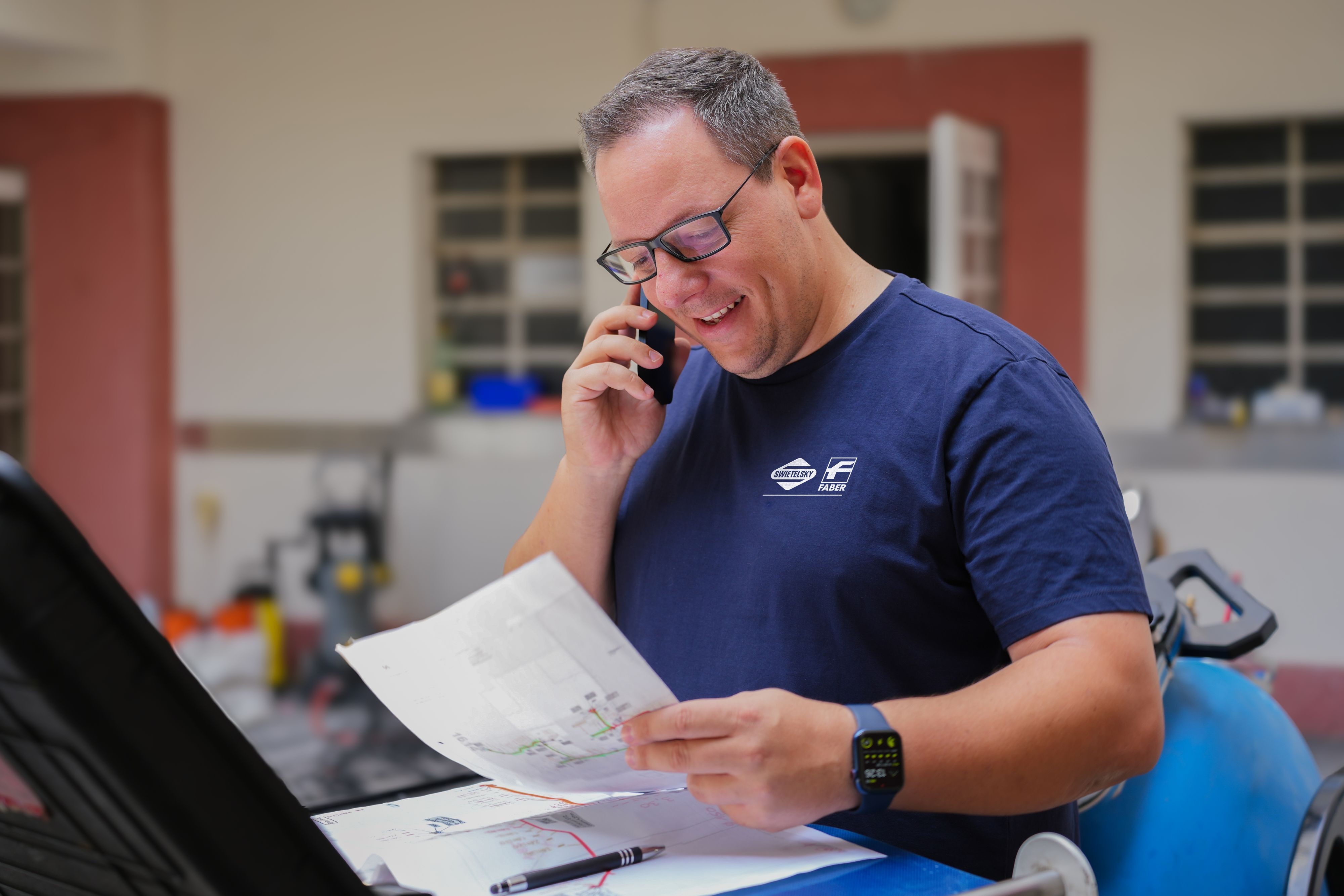 Ein Mann im marineblauen Shirt telefoniert und liest dabei ein Blatt neben einem Arbeitsplatz.