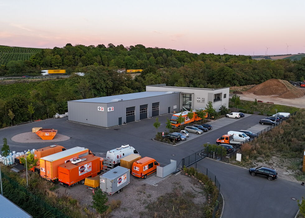 Luftbild eines Industriekomplexes mit weißen Gebäuden, orangen Maschinen und abgestellten Fahrzeugen bei Dämmerung.